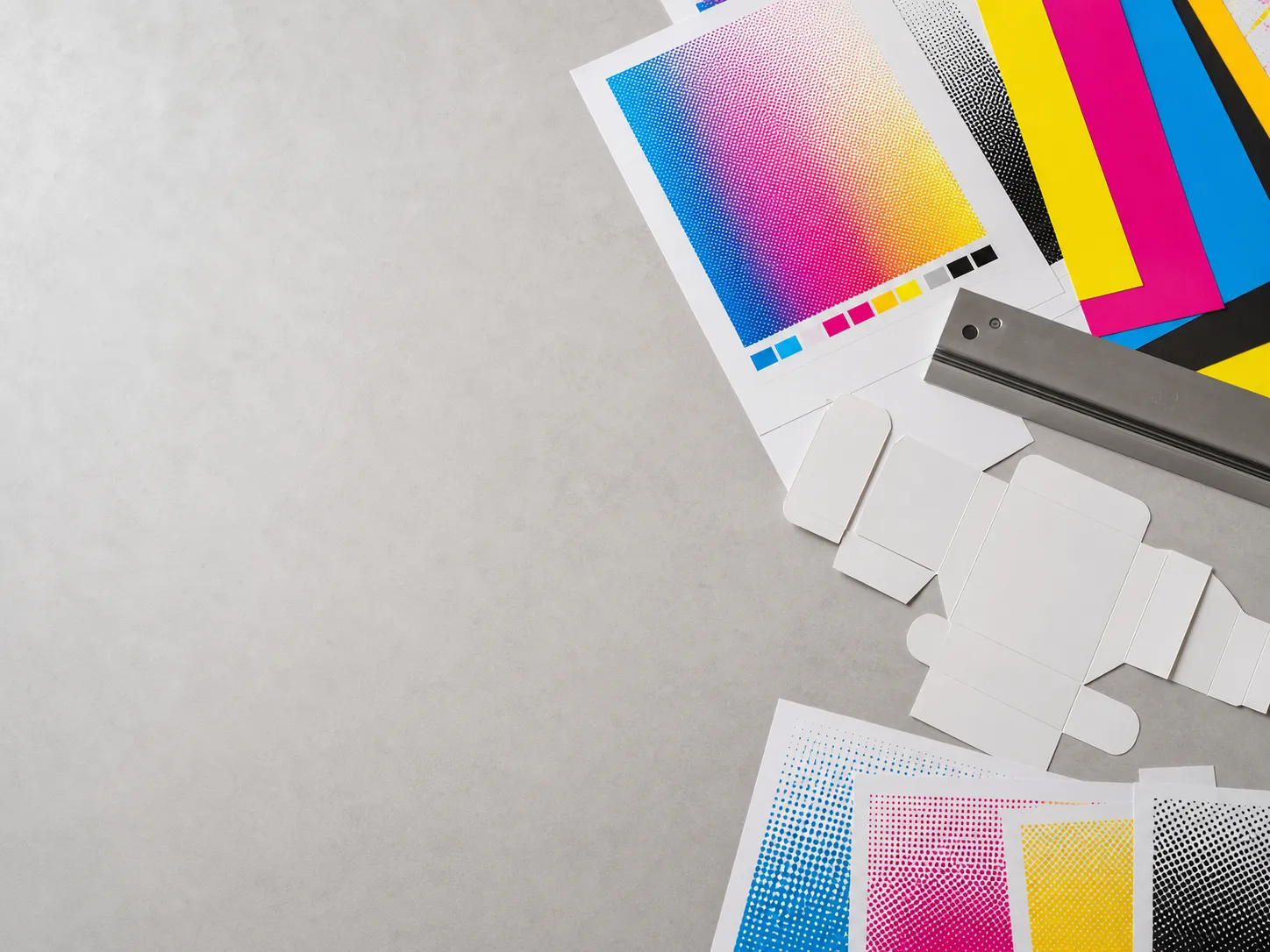 ලා අළු වැඩ මතුපිටක CMYK process sheets, හිස් packaging dieline හැඩ, halftone swatches සහ වානේ මිනුම් දණ්ඩක් දැක්වෙන, ඉහළින් බැලූ original print studio දසුනක්.
